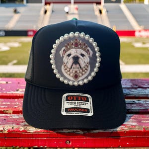 May include: A black trucker hat featuring a circular patch with a white bulldog wearing a gold crown and pearl necklace, surrounded by a border of white faux pearls. A rectangular sticker on the brim reads OTTO WORLD FAMOUS FOAM FRONT TRUCKER.