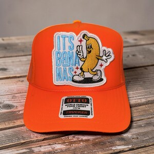 May include: Orange trucker hat with a patch featuring a cartoon banana character and the text "ITS BON NAS" in blue. The hat has a foam front and a durable snap closure. The brand "OTTO" is also visible.