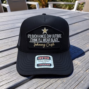 May include: Black trucker hat with a foam front and mesh back. The hat features a patch with the text "IT'S SUCH A NICE DAY OUTSIDE I THINK I'LL WEAR BLACK" and "Johnny Cash" in white and gold lettering. A gold star is above the text.