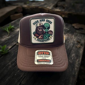 May include: Brown trucker hat with a patch featuring a Bigfoot and an alien with the text "HIDE AND SEEK CHAMPIONS". The hat has a brown foam front, a beige mesh back, and a snap closure.