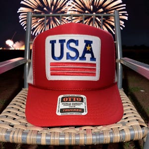 May include: Red trucker hat with a white mesh back and a white patch featuring the word "USA" in blue letters and a gold star. The hat has a curved brim and a "OTTO" label.
