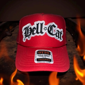 May include: A red trucker hat with the text "Hell Cat" in a stylized font. The hat has a mesh back and a snap closure. The front of the hat features a white and black embroidered design.