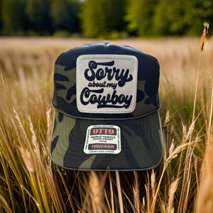 May include: A camouflage trucker hat with a white patch that says "Sorry about my Cowboy". The hat has a black mesh back and a snap closure.