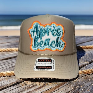 May include: A tan trucker hat with a mesh back and a curved brim. The front of the hat features a patch with the words "Après Beach" in turquoise script with orange and blue accents. The hat has a snap closure.