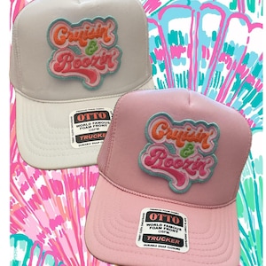 May include: Two trucker hats with a mesh back. The hats are pink and gray with a white, orange, and green embroidered patch that says "Cruisin' & Boozin'". The hats have a label that says "Otto World Famous Foam Front OSFM Trucker Durable Snap Closure".