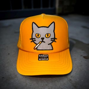 May include: Yellow trucker hat with a gray cat graphic smoking a cigarette. The cat has yellow eyes and black whiskers. The hat has a mesh back and a curved brim. The brand logo is visible on the front.