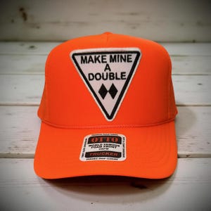 May include: Bright orange trucker hat with a white triangle patch featuring the text "MAKE MINE A DOUBLE" and two black diamond shapes. The hat has a foam front and a durable snap closure.