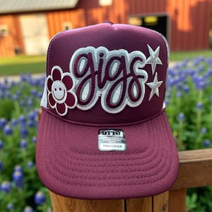 May include: Burgundy trucker hat with a white mesh back. The front of the hat features a white and silver embroidered "gigs" with a white daisy with a smiley face and two silver stars.