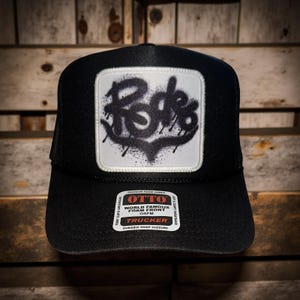 May include: Black trucker hat with a white square patch featuring the word "Rode" in a graffiti-style design. The hat has a durable snap closure and a foam front. The hat is made of soft and comfy sweatband material.