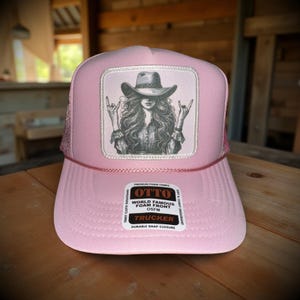 May include: Pink trucker hat with a patch featuring a woman in a cowboy hat making a rock-and-roll hand gesture. The hat has a mesh back and a curved brim. The patch reads "OTTO WORLD FAMOUS FOAM FRONT TRUCKER DURABLE SNAP CLOSURE".
