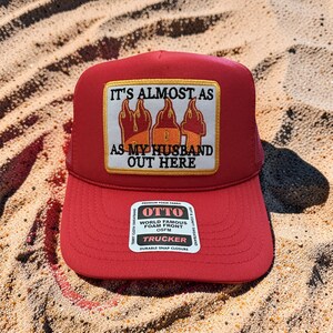 May include: Red trucker hat with a foam front and a mesh back. The hat features a rectangular patch with the text "IT'S ALMOST AS AS MY HUSBAND OUT HERE" above a graphic of three stylized flames. The hat has a durable snap closure.