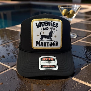 May include: Black trucker hat with a square patch featuring the text "WEENIES + AND MARTINIS" and a dachshund dog illustration. The patch has a gold border. The hat has a mesh back and a curved brim.