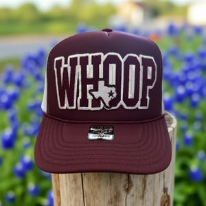 May include: Maroon trucker hat with the word "WHOOP" in white block letters. The Texas state outline with a star is in the center of the word. The hat has a white mesh back and a maroon brim. The hat is sitting on a wooden post.