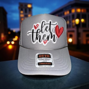 May include: Gray trucker hat with the words "let them" in black script and red heart accents. The hat has a mesh back and a snap closure. The brand tag reads "OTTO World Famous Foam Front OSFM Trucker".