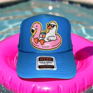 May include: A blue trucker hat with a mesh back and a flamingo and duck graphic on the front. The flamingo and duck are in a pink inflatable ring, with the duck wearing sunglasses and holding a drink. The hat has a snap closure.