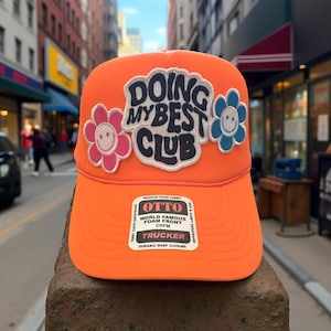 May include: Orange trucker hat with a black, white, and blue patch that says "Doing My Best Club". The patch is surrounded by two embroidered flowers with smiley faces. The hat has a black label that says "Otto World Famous Foam Front OSFM Trucker Durable Snap Closure".