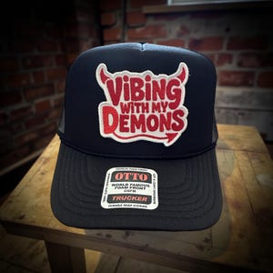 May include: Black trucker hat with the text "Vibing With My Demons" in red and white. The hat has a mesh back and a curved brim. The hat also has a tag that says "OTTO World Famous Foam Front".