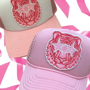 May include: Two pink and white trucker hats with a pink tiger face with star-shaped eyes embroidered on the front.