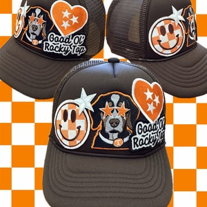 May include: Brown trucker hat with a variety of patches, including a smiley face, a dog with stars over its eyes, a heart with stars, and a patch that says "Good Ol' Rocky Top".