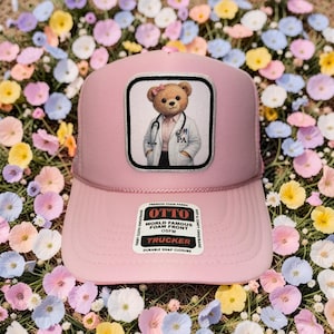 May include: Pink trucker hat with a patch featuring a teddy bear dressed as a PA. The hat has a pink brim and mesh backing. The patch has the text "OTTO" and "TRUCKER".