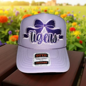 May include: A lavender trucker hat with a purple bow and the word "Tigers" embroidered on the front. The hat has a mesh back and a snap closure. The hat has a label that says "OTTO WORLD FAMOUS FOAM FRONT".