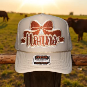 May include: A beige trucker hat with a brown bow and the word "horns" embroidered on the front. The hat has a mesh back and a snap closure. The hat is sitting on a wooden fence post with a field and cows in the background.