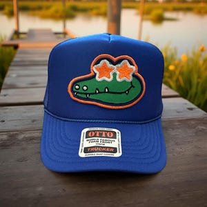 May include: A blue trucker hat with a green alligator with orange eyes and two orange stars on top. The hat has a black mesh back and a white label that says "OTTO WORLD FAMOUS FOAM FRONT OSFM TRUCKER DURABLE SNAP CLOSURE".