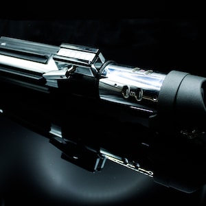 May include: A silver and black lightsaber with a black handle and a silver blade. The lightsaber is on a black surface.