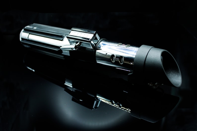 May include: A silver and black lightsaber with a black handle and a silver blade. The lightsaber is on a black surface.
