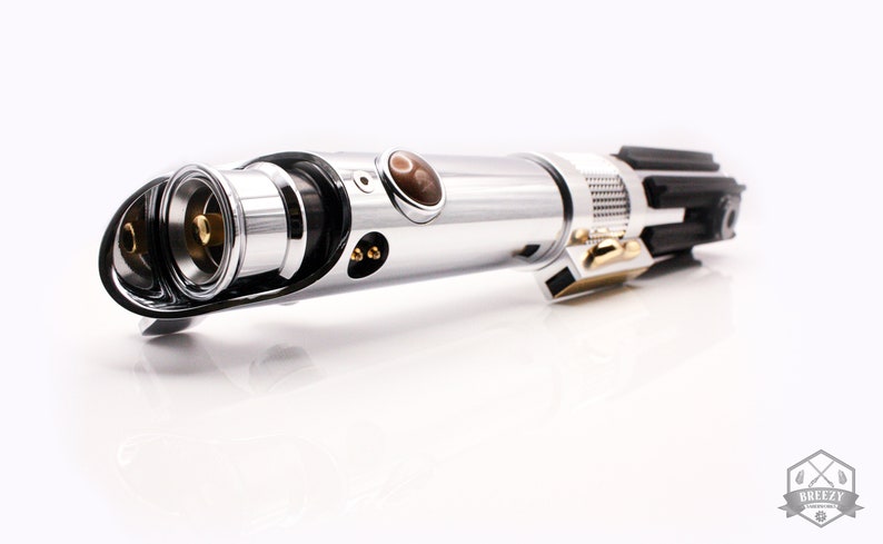 May include: A silver lightsaber with a black handle and a gold accent. The lightsaber has a brown crystal and a black and silver emitter.