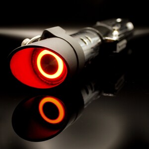 May include: A black lightsaber with a red glowing blade. The lightsaber is on a black surface.