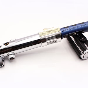 May include: A silver lightsaber with a gold accent and a blue and black lightsaber hilt. The hilt has the text "NEW YORK CITY" on it.