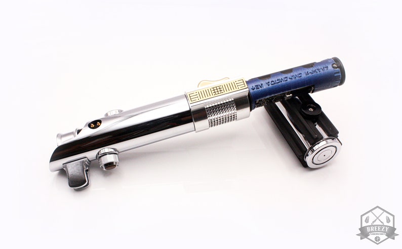 May include: A silver lightsaber with a gold accent and a blue and black lightsaber hilt. The hilt has the text "NEW YORK CITY" on it.