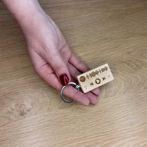 Op de afbeelding: Een houten sleutelhanger met een Spotify-logo en een barcode-achtig ontwerp. De sleutelhanger heeft ook play-, pauze- en skipknoppen.