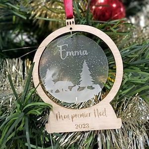 May include: A wooden Christmas ornament with a frosted glass globe featuring a snowy scene with trees and bears. The globe is inscribed with the name "Emma" and the text "Mon premier Noel 2023".