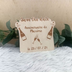 Può includere: Carta di legno "Save the date" con un design di bottiglia di champagne e bicchieri. Il testo recita "Save the date, Anniversaire de Maxime, le 23/07/24".