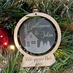 May include: A wooden Christmas ornament with a clear acrylic insert. The insert features a house with a chimney, two trees, and the text "Guillaume & Julia". The ornament also has the text "Notre premier Noel 2023" on the bottom.
