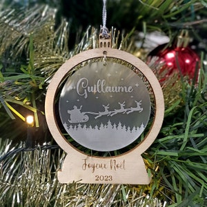 May include: A wooden Christmas ornament with a clear glass globe featuring a Santa Claus silhouette in a sleigh pulled by reindeer. The globe has the name "Guillaume" and the text "Joyeux Noel 2023" inscribed on the wood.