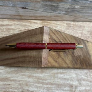 以下が含まれることがあります： 金色のアクセントが付いた赤い木製のペン。ペンは木製の表面の上に置かれています