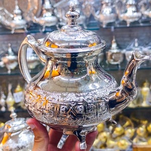 Peut inclure: Une théière en métal argenté poli présentant un motif géométrique et floral en relief complexe sur tout le corps. La théière est dotée d'une anse incurvée, d'un long bec verseur, d'un couvercle amovible avec un petit bouton et repose sur trois pieds.