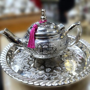 Könnte beinhalten: Verzierte silberne Teekanne mit passendem Tablett. Die Teekanne hat aufwendige eingravierte Muster und eine rosa Quaste auf dem Deckel. Das Tablett hat einen dekorativen, geriffelten Rand. Ein klassisches Teeservice.