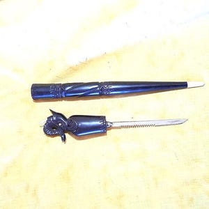 May include: A black, carved letter opener with a matching sheath. The handle of the letter opener features a detailed ram's head design. The sheath is long and tapered, with carved details. The blade is serrated.