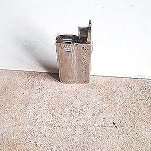 May include: A vintage, metallic lighter with a rectangular body and a hinged lid. The lighter has a textured surface and a small, vertical lever. The design is simple and functional, with a classic aesthetic.