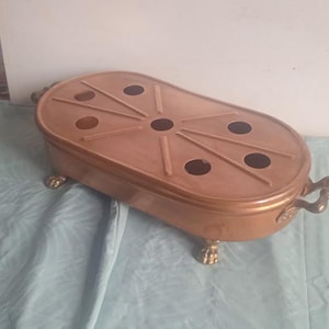Peut inclure: Un plat de service ovale en cuivre avec six découpes circulaires et quatre pieds décoratifs. Le plat a une finition dorée et est conçu pour contenir des aliments chauds.