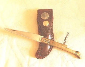 May include: A folding pocket knife with a silver-toned blade, a light-colored handle featuring an engraved figure, and a metal corkscrew attachment. The knife is positioned over a brown leather sheath with braided edges and a metal snap closure.