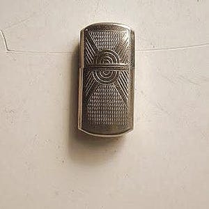 May include: A silver lighter with a geometric design and a circular pattern in the centre.