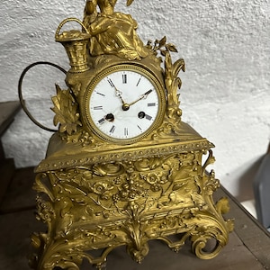 Puede incluir: Un reloj de chimenea dorado con una base ornamentada y una figura de una mujer sosteniendo una cesta en la parte superior. La esfera del reloj tiene números romanos y un fondo blanco.
