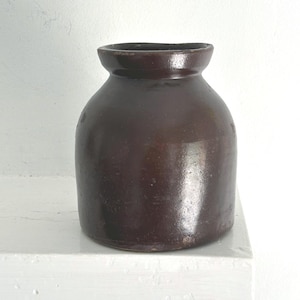 May include: A small, dark brown ceramic crock with a smooth, glazed finish. The vessel features a wide, rounded body, a short neck, and a thick, slightly flared rim. The surface has a reflective sheen and a uniform, deep brown color throughout.