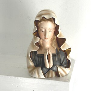 May include: A ceramic bust of a figure with closed eyes and hands pressed together in prayer. The figure wears a dark grey garment and a draped, cream-colored head covering with gold-toned interior lining, featuring soft, rounded facial features.