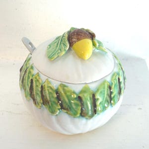 May include: A white ceramic sugar bowl with a lid, decorated with green leaves and a brown and yellow acorn. A small spoon rests on the side. The bowl has a bulbous shape and is likely for kitchen use.